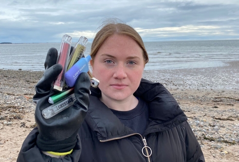 Woman on beach holding up a handful of discarded disposable vapes, wearing a big jacket and looking stern. 