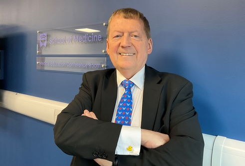 Professor Tom MacDonald stands with arms folded in front of a blue background.