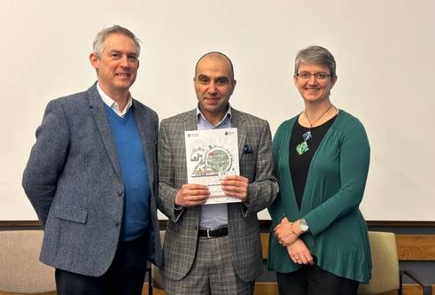 Three people grouped together. A male in the centre holds a booklet and there is a male on his left and a female on his right. All are smartly dressed and smiling