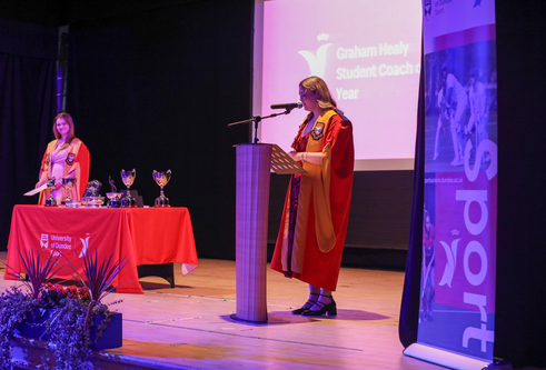 A women in a robe stands in front of a pedestal speaking in a microphone. Another person stands behind a table covered in red cloth.