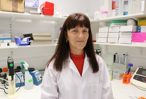 Woman with long dark hair, wearing white lab coat, standing in a lab with shelves behind her with lots of science materials on them.