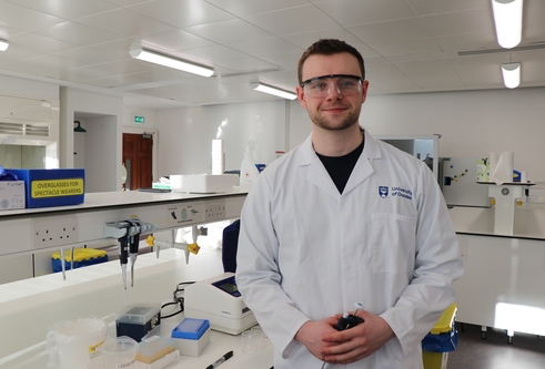 Young man wearing white lab coat and protective eye wear, standing in a science lab with science equipment around him