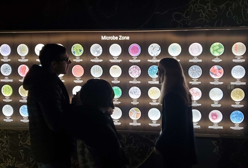 Professor Sarah Coulthurst speaking to members of the public in front of a wall displaying lots of microbes at Dundee Science Centre