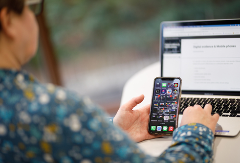 A person sitting at a table with a mobile phone in one hand and a laptop on the table