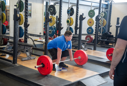 A boy squats over a set of weights, ready to lift. There are lots of weights stored on racks around the room behind him.