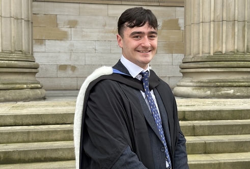 Young male with dark hair wearing graduation gown and hood, with suit and tie underneath