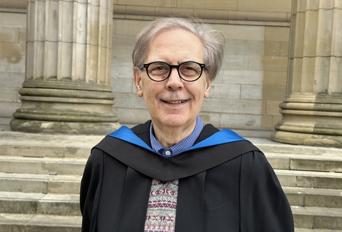 Man aged 75 with greying hair parted at the side and down to eye level, wearing glasses and graduation gown and hood and smiling at the camera