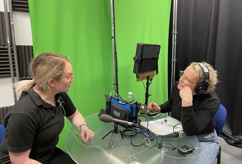 Two women sitting down at a table facing each other and talking, both wearing headphones. On the table are microphones pointing at each of them and lots of audio recording equipment.