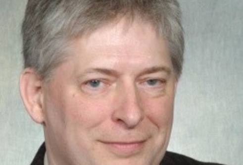 Man with grey hair in a shirt, tie and blazer against a grey back ground with a slight smile