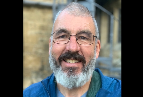 man in a blue shirt and grey jumper is stood in a lab smiling at the camera