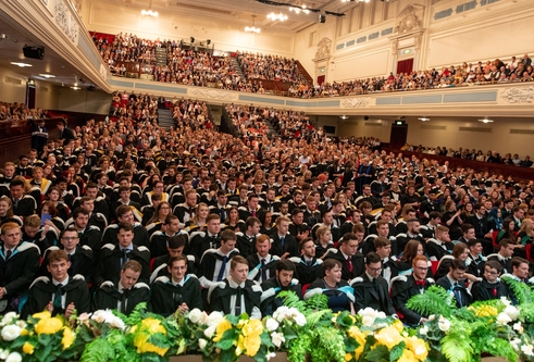 Hundreds of students sitting together in a large hall for their graduation ceremony. There is a upper level for their families.  