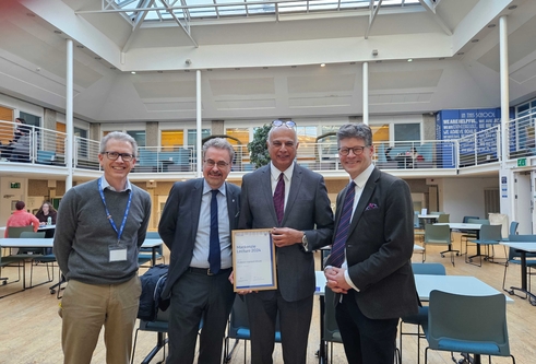 Four men wearing suits stand together holding a framed certificate for the Mackenzie Lecture 2024