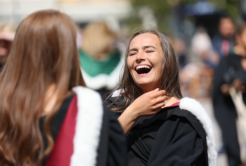 Two women in graduation gowns with red hoods lined with white fur. One stands with her back to us, the other faces us and is laughing.