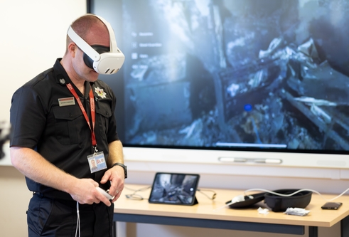 Man wearing fire fighter uniform and virtual reality headset over his eyes, with a large screen behind him showing a fire scene