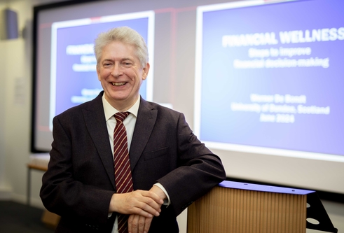 A man wearing a suit with a red tie leans against a podium, there are screens behind him showing a presentation.