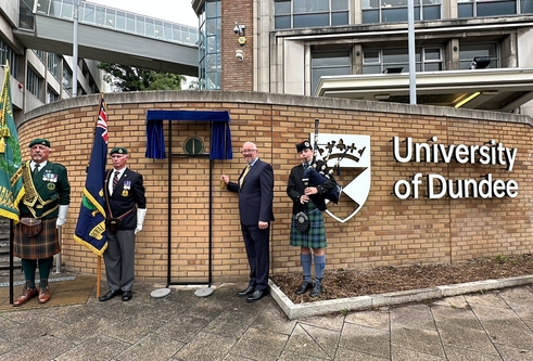 Two men in formal wear holding flags, one man in a suit standing next to a plaque and a piper