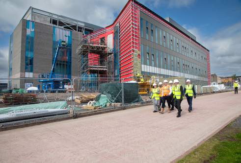 A group of people in high-vis vests walking in front of a building site. There is a very large building in the site.