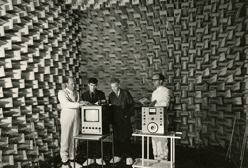 Four men stand inside an Anechoic Chamber. They are looking at two pieces of equipment, one a monitor, the other some sort of measurement device