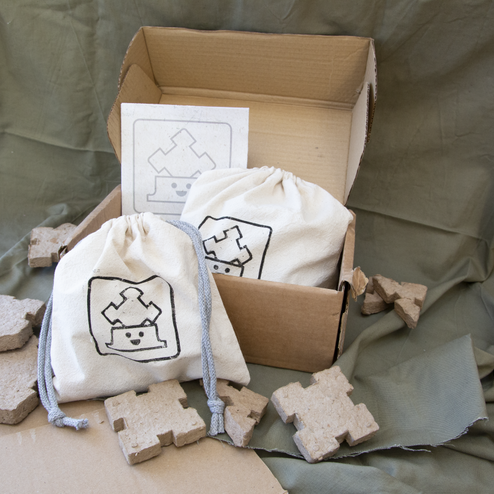 A cardboard box containing a filled drawstring bag and an instruction booklet is situated on a green backdrop. Another drawstring bag printed with the Ecoform logo is next to the cardboard box, which is surrounded with paper pulp shapes.