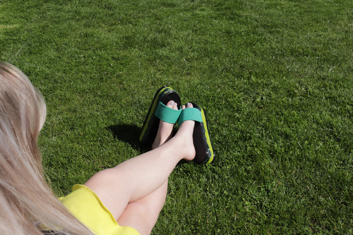 Back of a girls head with legs and feet showing. Wearing green and black sliders. Girl sitting on grass.